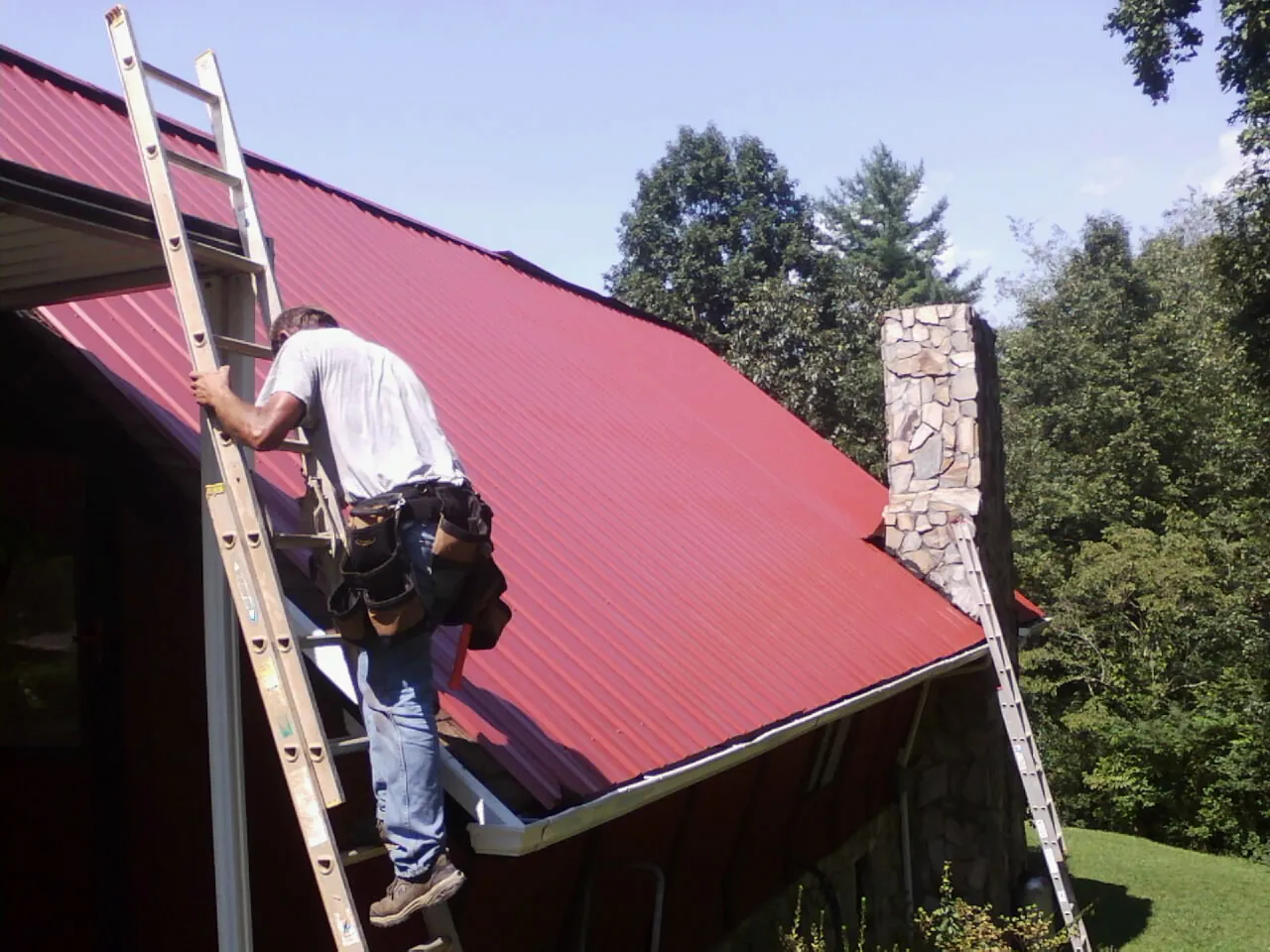 Roofer on a ladder beside a red metal roof and stone chimney — Soffit & Fascia Repair by Roofing Brothers in West Warren